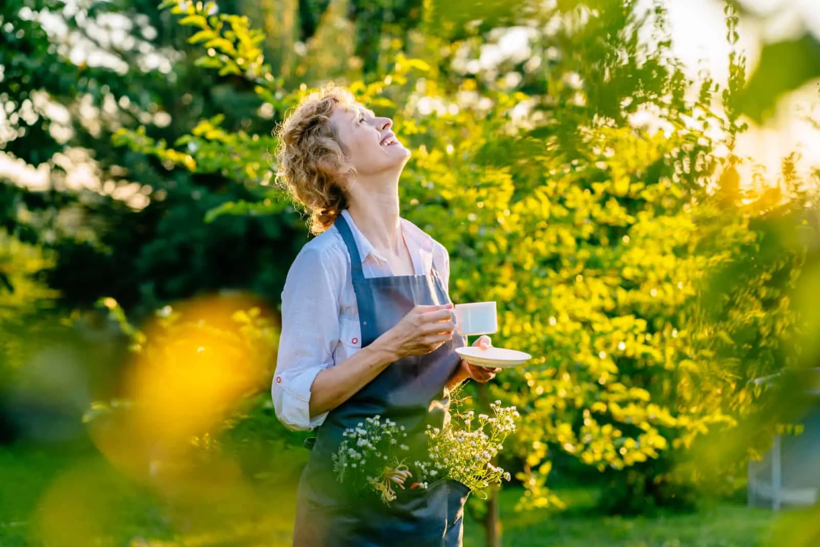 Happy serene woman gardener in grey apron drinking coffee in garden outdoor.