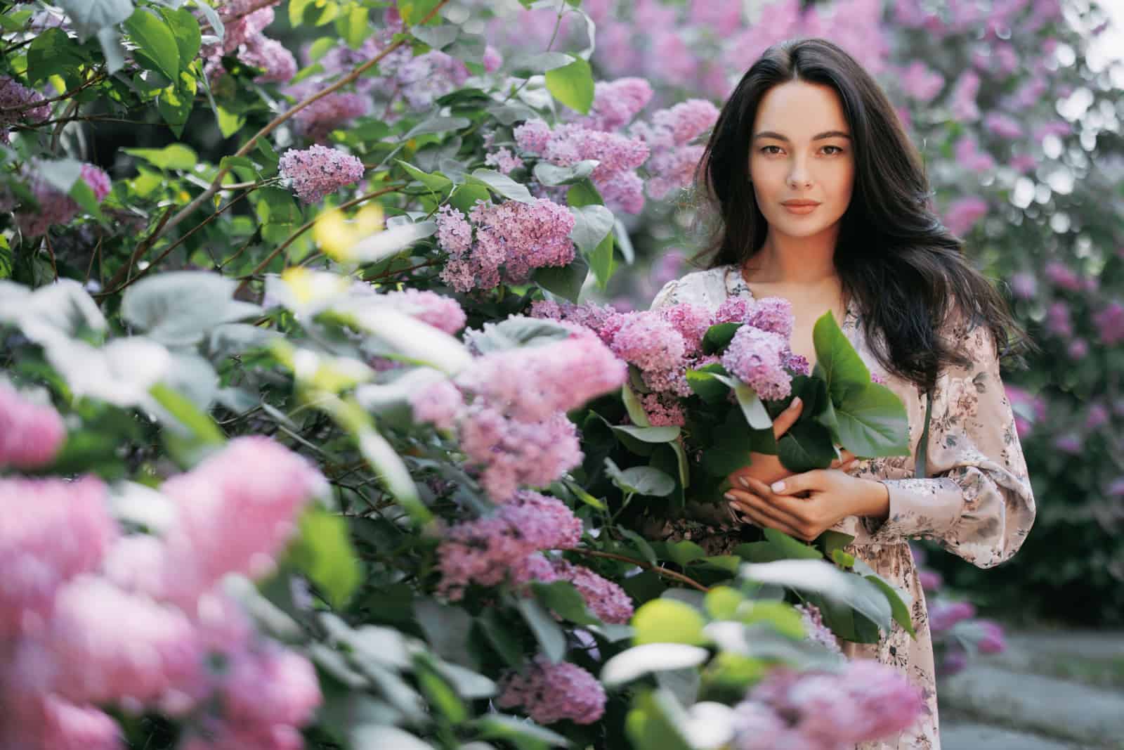 Young pretty brunette woman walks in garden with bouquet in her hands and enjoys by blooming lilac shrubs.