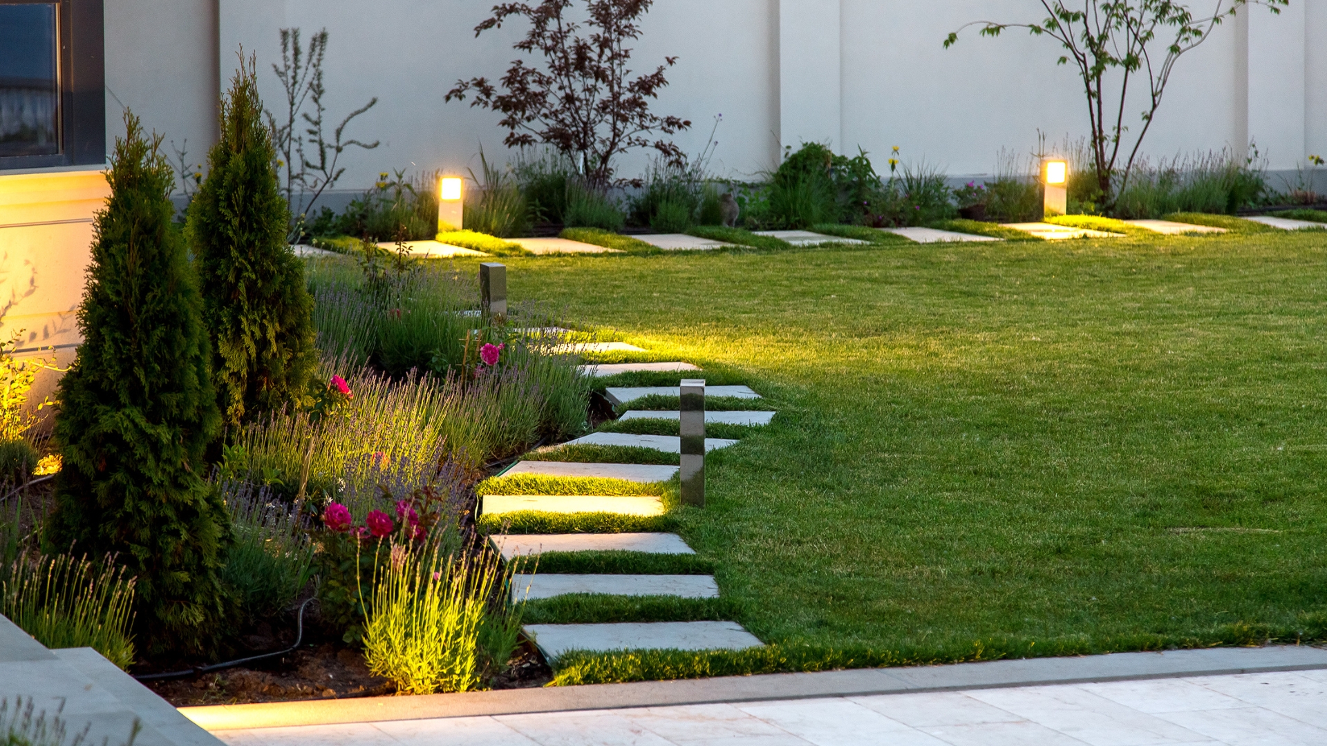 backyard of the mansion with a flowerbed and a lawn of green grass with a marble walkway of square tiles in the evening with a garden lighting with decorative ground lamps illuminating a warm light.