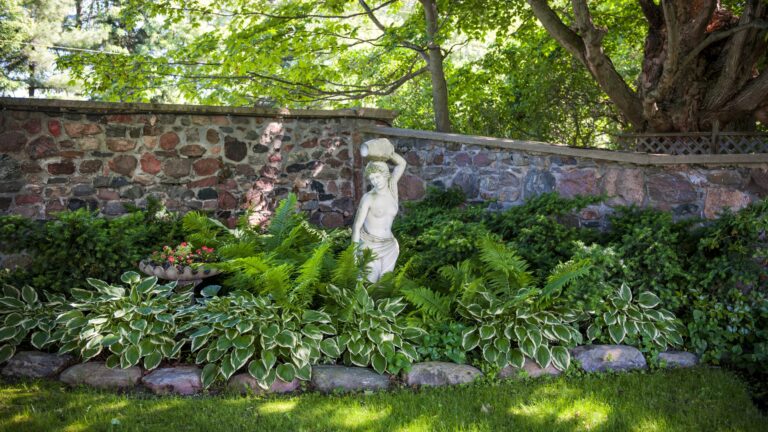 Lush green summer garden with perennial plants and statue near stone wall
