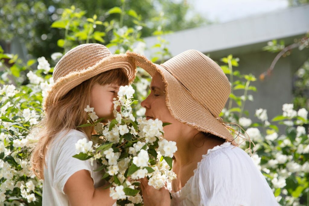 mom and little daughter wearing straw hats embrace amid flowering jasmine