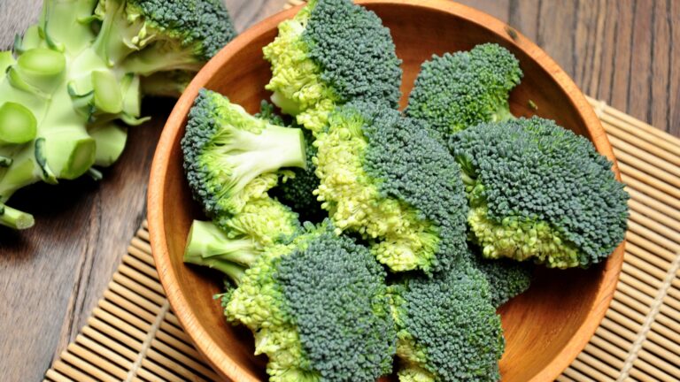 Close-up of a wooden bowl filled with vibrant green broccoli florets. The broccoli is surrounded by a few whole broccoli stalks on a wooden surface.